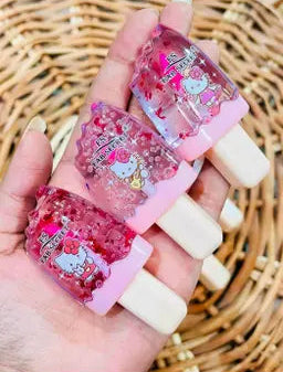 Three pink Hello Kitty gel popsicle-style nail wraps held in a hand, featuring pink glitter and Hello Kitty illustrations, with a woven straw background.