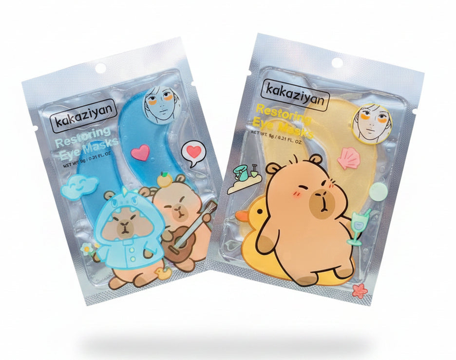 Two packages of kakaziyan Restoring Eye Masks held in a hand against a background of green foliage. One package contains a blue mask and the other contains a yellow mask, both featuring cute cartoon capybara illustrations.