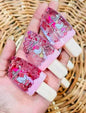 Three pink Hello Kitty gel popsicle-style nail wraps held in a hand, featuring pink glitter and Hello Kitty illustrations, with a woven straw background.