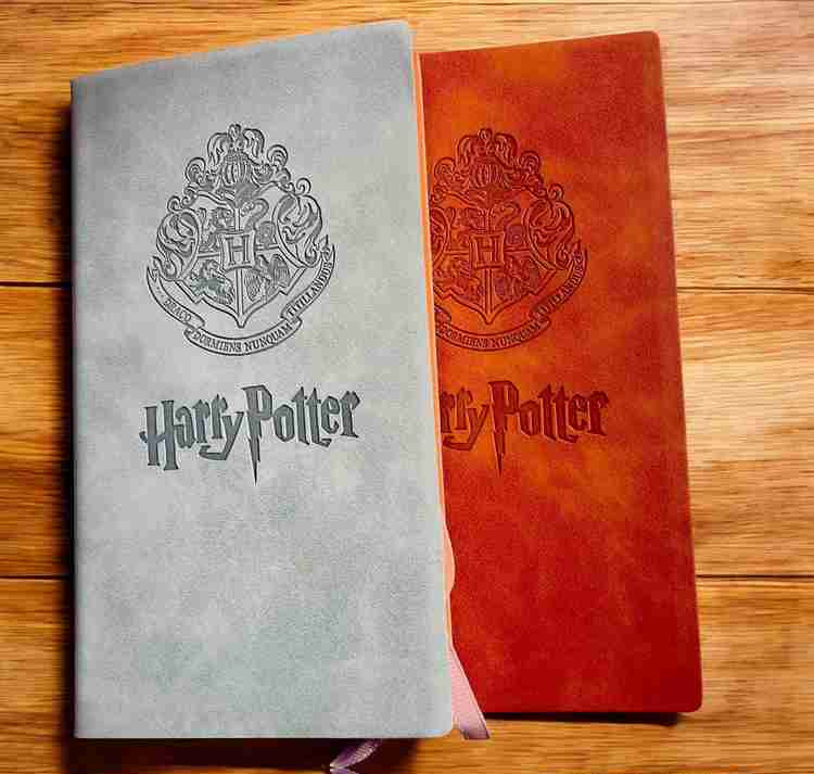 Two Harry Potter hardcover notebooks on a wooden surface, one in textured light gray with a dark gray crest and title, and one in textured reddish-orange with a dark red crest and title. A pink ribbon bookmark is visible from the gray notebook.