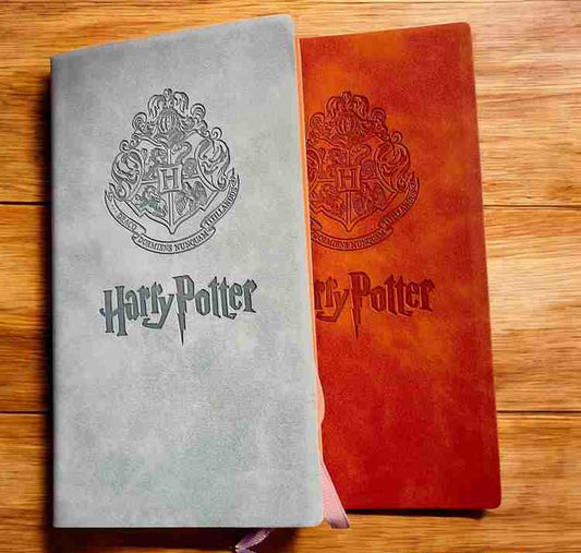 Two Harry Potter hardcover notebooks on a wooden surface, one in textured light gray with a dark gray crest and title, and one in textured reddish-orange with a dark red crest and title. A pink ribbon bookmark is visible from the gray notebook.