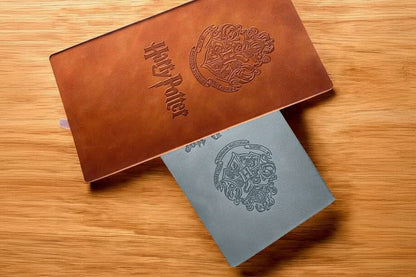 A Harry Potter themed brown leather-like box with embossed crest and logo, partially slid open to reveal a silver-gray insert card with a gray embossed crest, placed on a wooden surface.